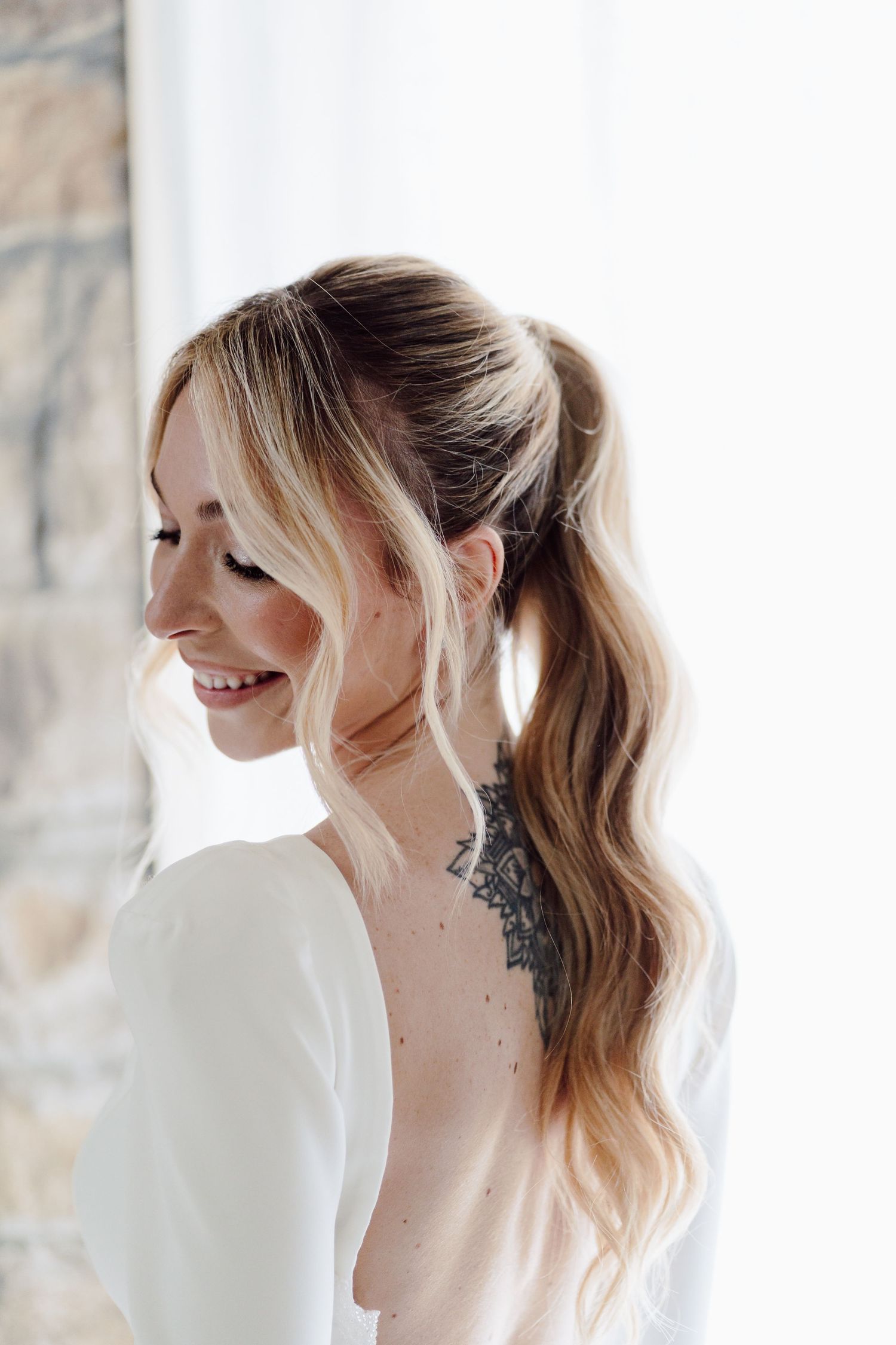 Bride with ponytail hairstyle and back tattoo smiling in white open-back wedding dress.