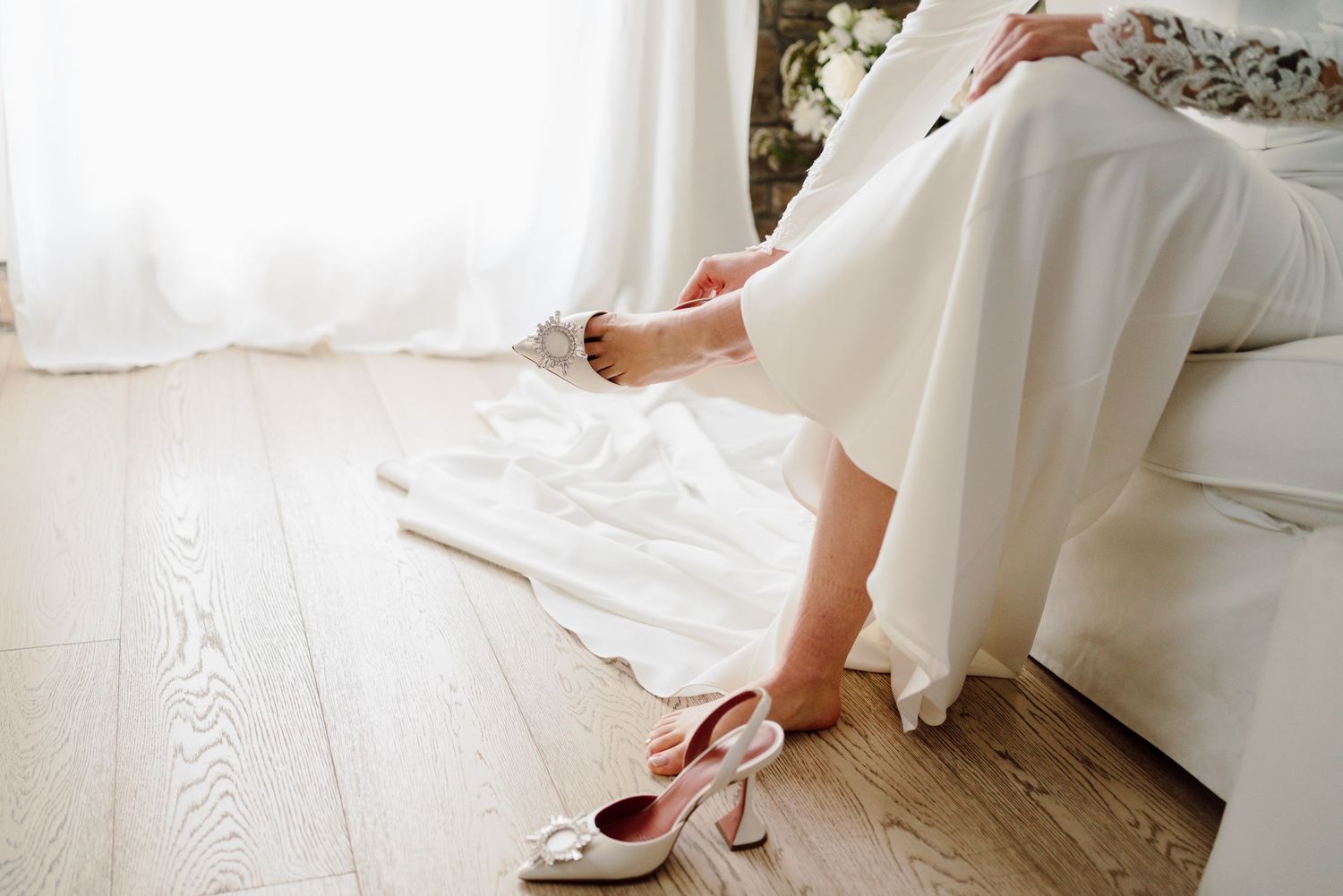 Bride in white wedding dress sitting on bed putting on pink heeled bridal shoes.