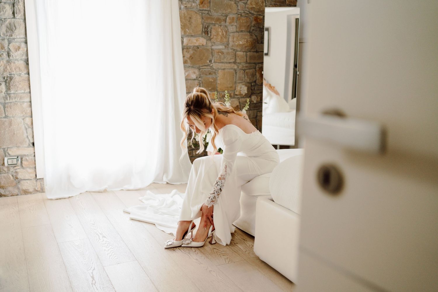 Bride in lace wedding gown sitting on bed putting on elegant high heel bridal shoes.