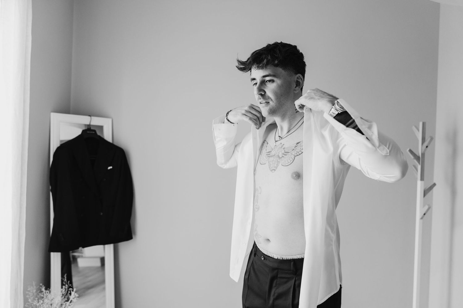 Groom getting dressed in white shirt with chest tattoo visible in black and white photo.