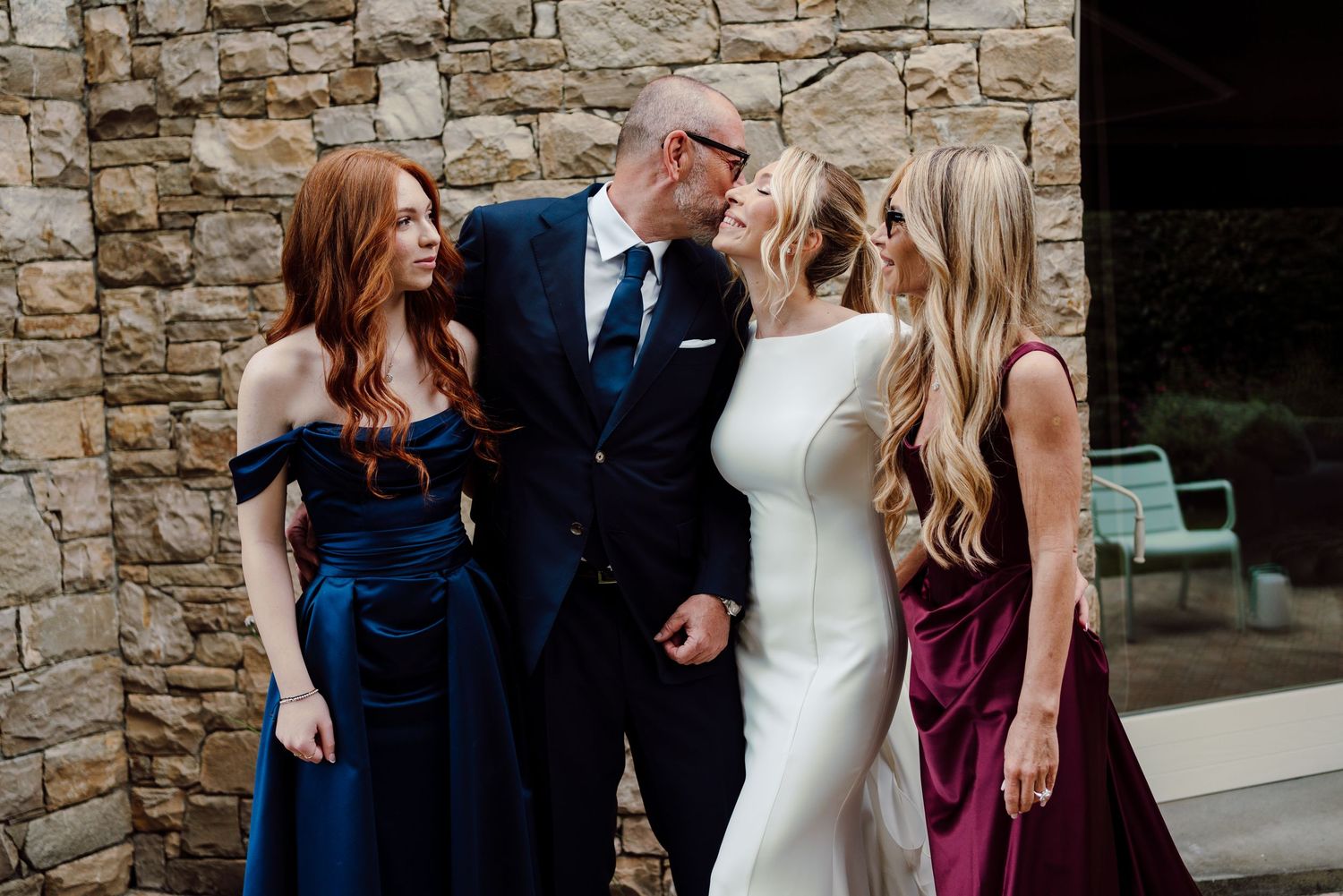 Bride in white gown with bridesmaids in navy and burgundy dresses beside a man in a suit at a wedding.