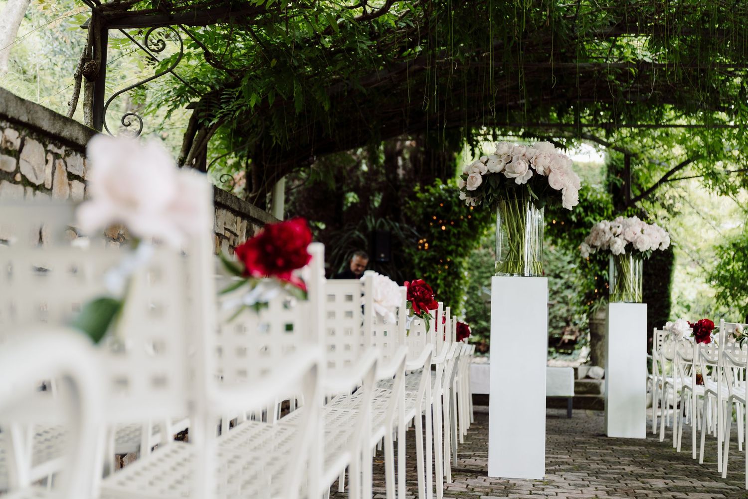 Elegant outdoor wedding ceremony aisle with white chairs and floral arrangements under a canopy of trees.