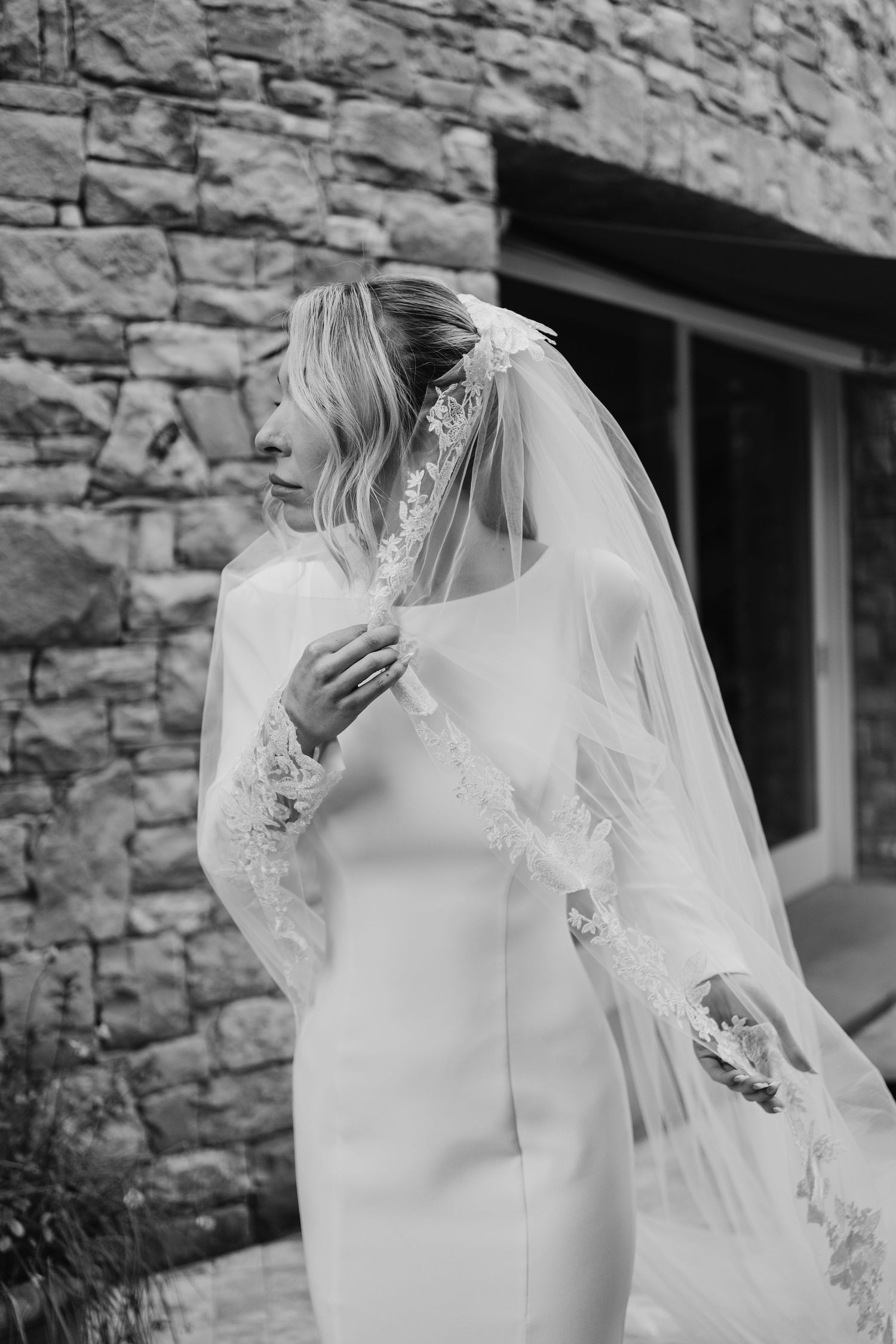 Black and white portrait of a bride in a white gown and lace veil standing by a stone wall.