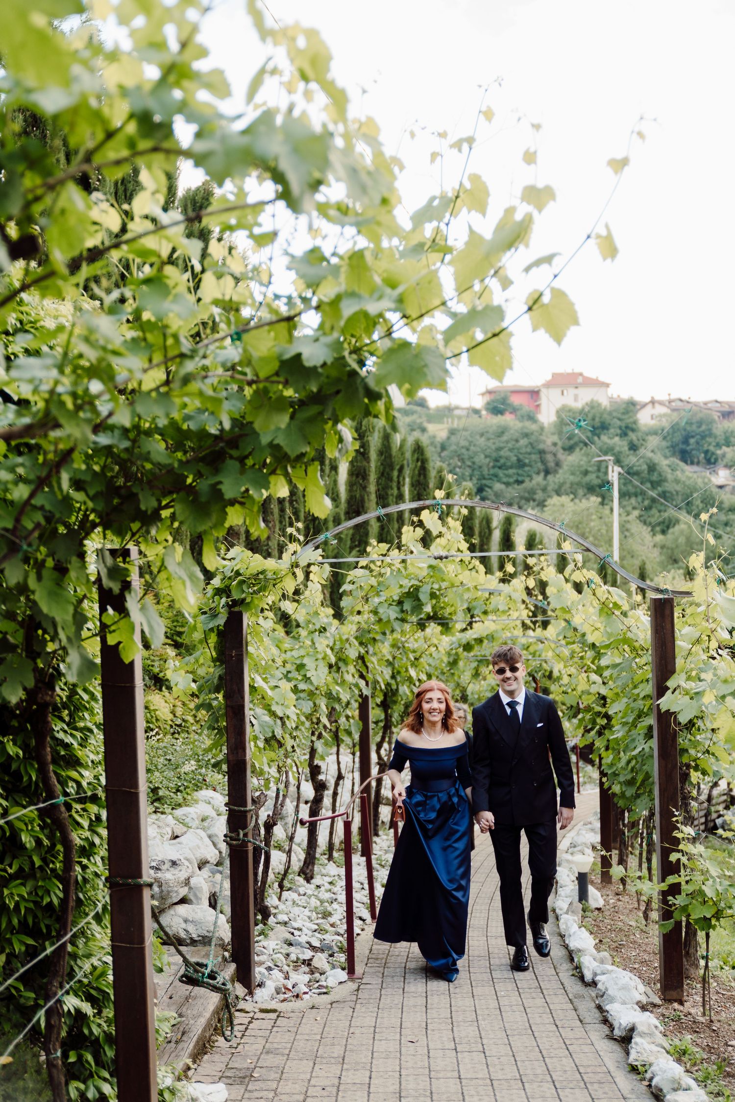 Couple walking hand in hand through a lush vineyard pathway in formal wedding attire.