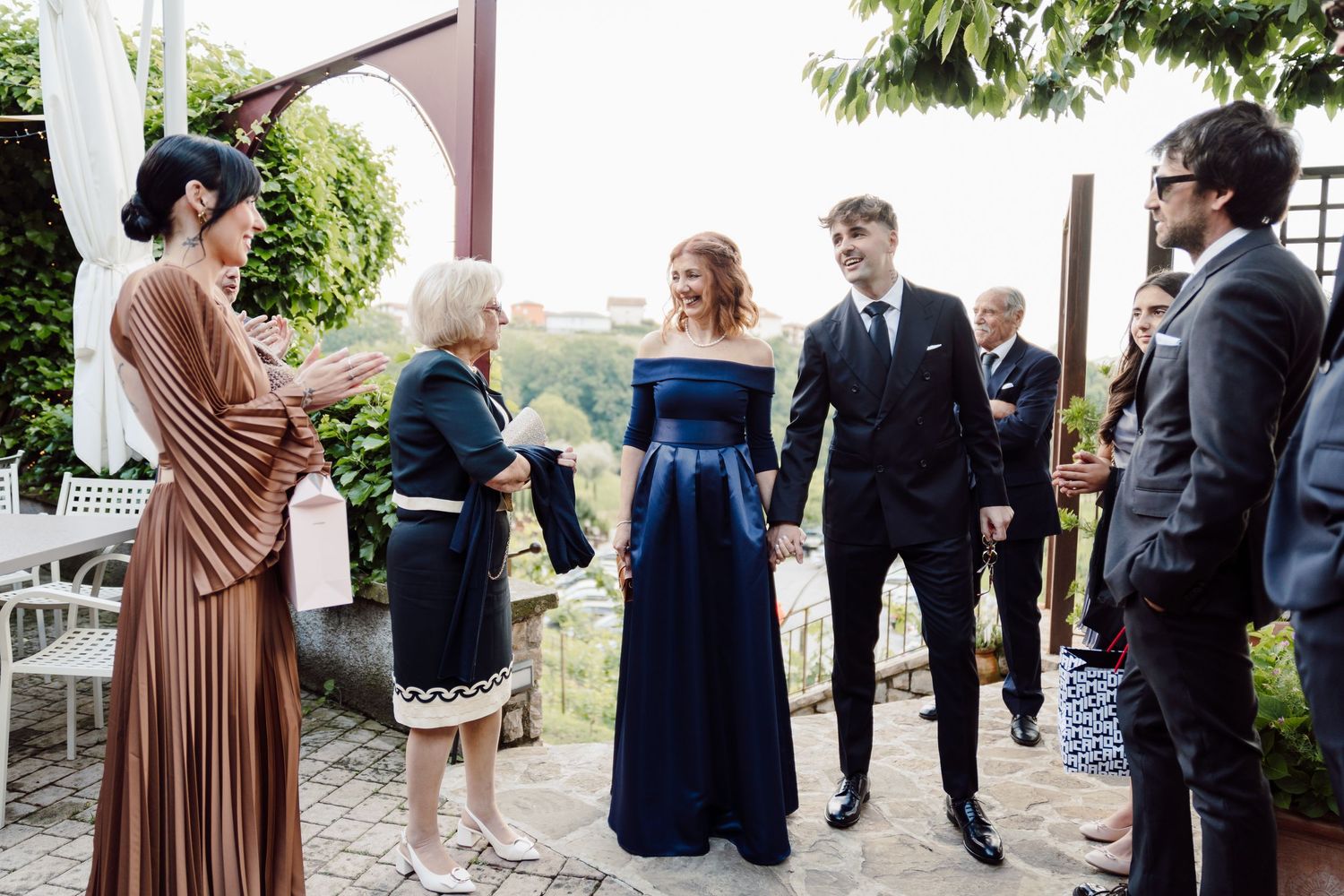 Guests greeting a couple in formal attire at an outdoor wedding reception in a garden setting.