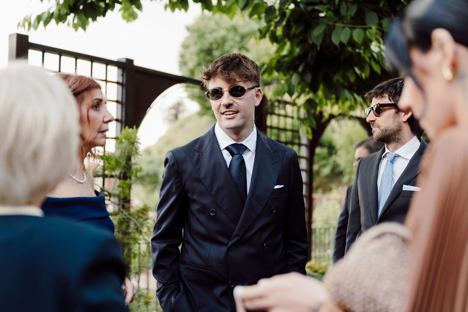 Stylish groom in a navy double-breasted suit and sunglasses socializing with wedding guests outdoors.