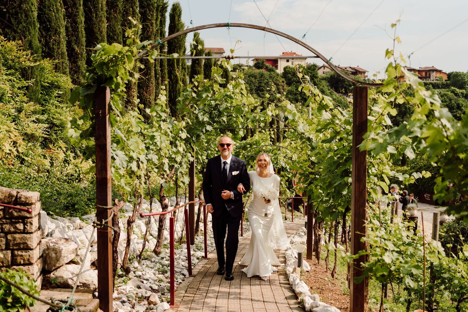Bride in white gown and veil walking through a vineyard arch with an escort on her wedding day.