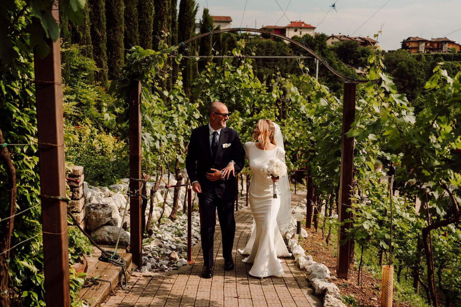 Bride and escort sharing a tender moment among lush vineyard rows on a sunny wedding day.