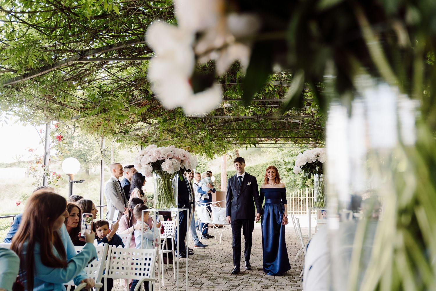 Groom escorts elegantly dressed woman in navy gown down outdoor wedding ceremony aisle.