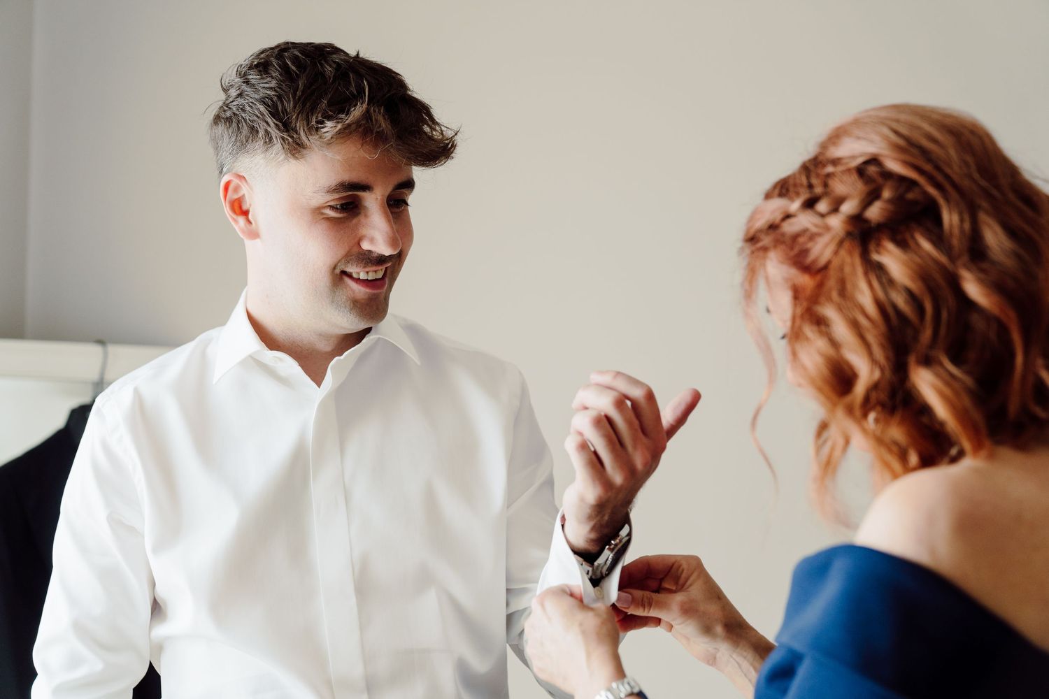 Smiling groom in white shirt having cufflinks fastened by woman in blue dress.