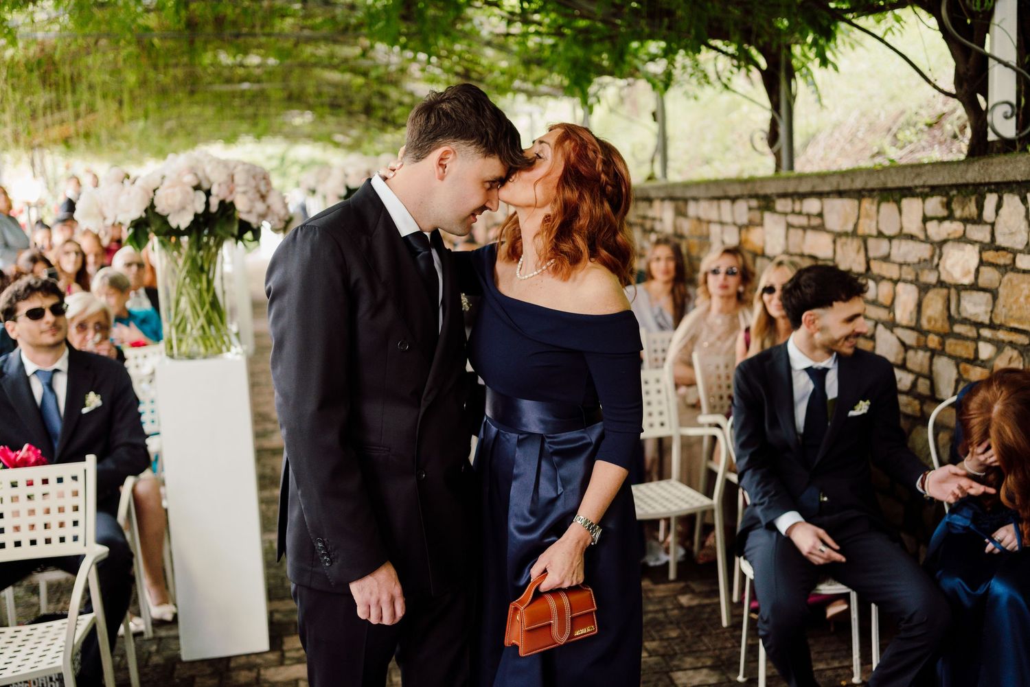 Groom shares a tender kiss with a woman in a navy off-shoulder dress during wedding ceremony.