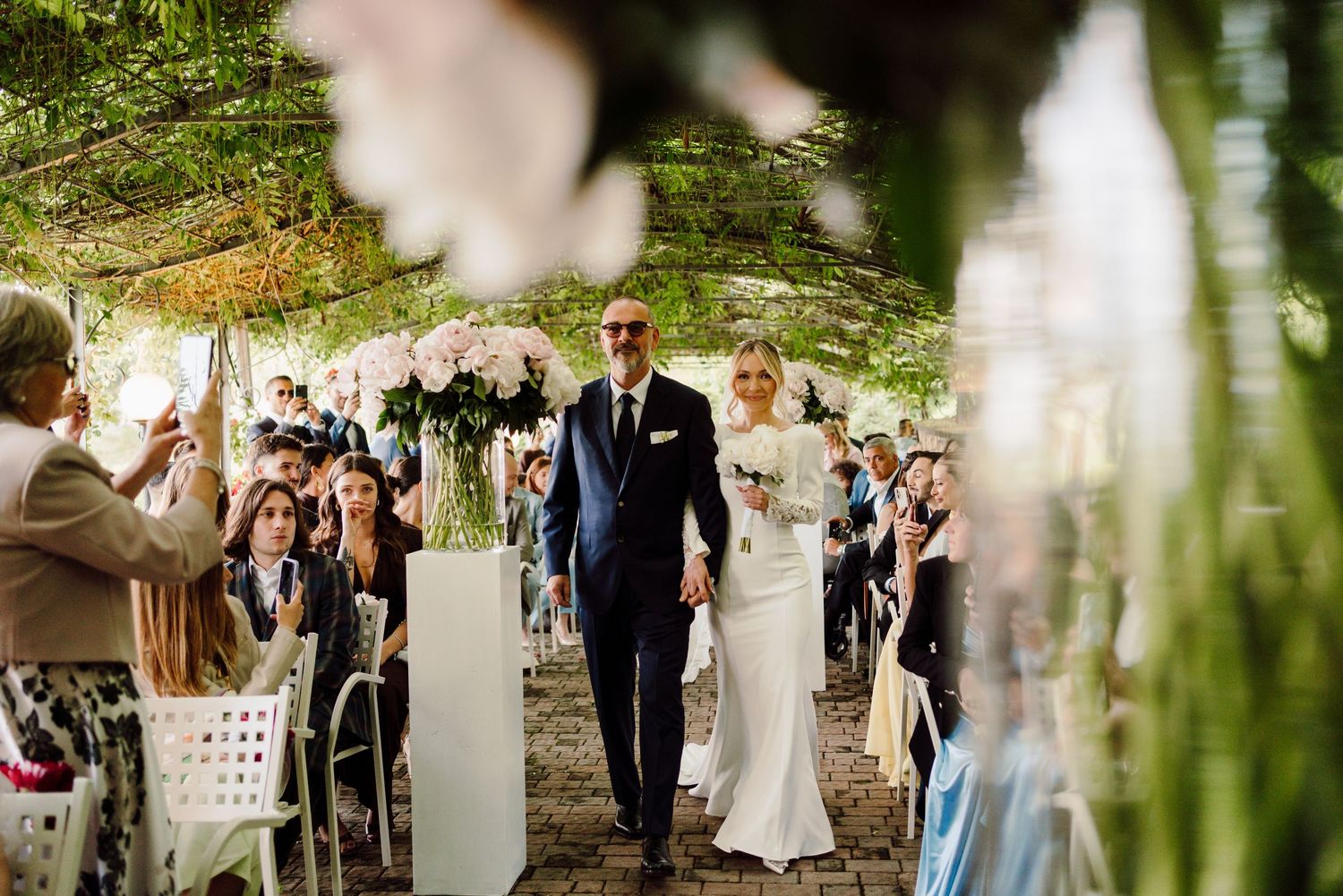 Father walks bride in elegant white gown down the aisle at an outdoor garden wedding ceremony.