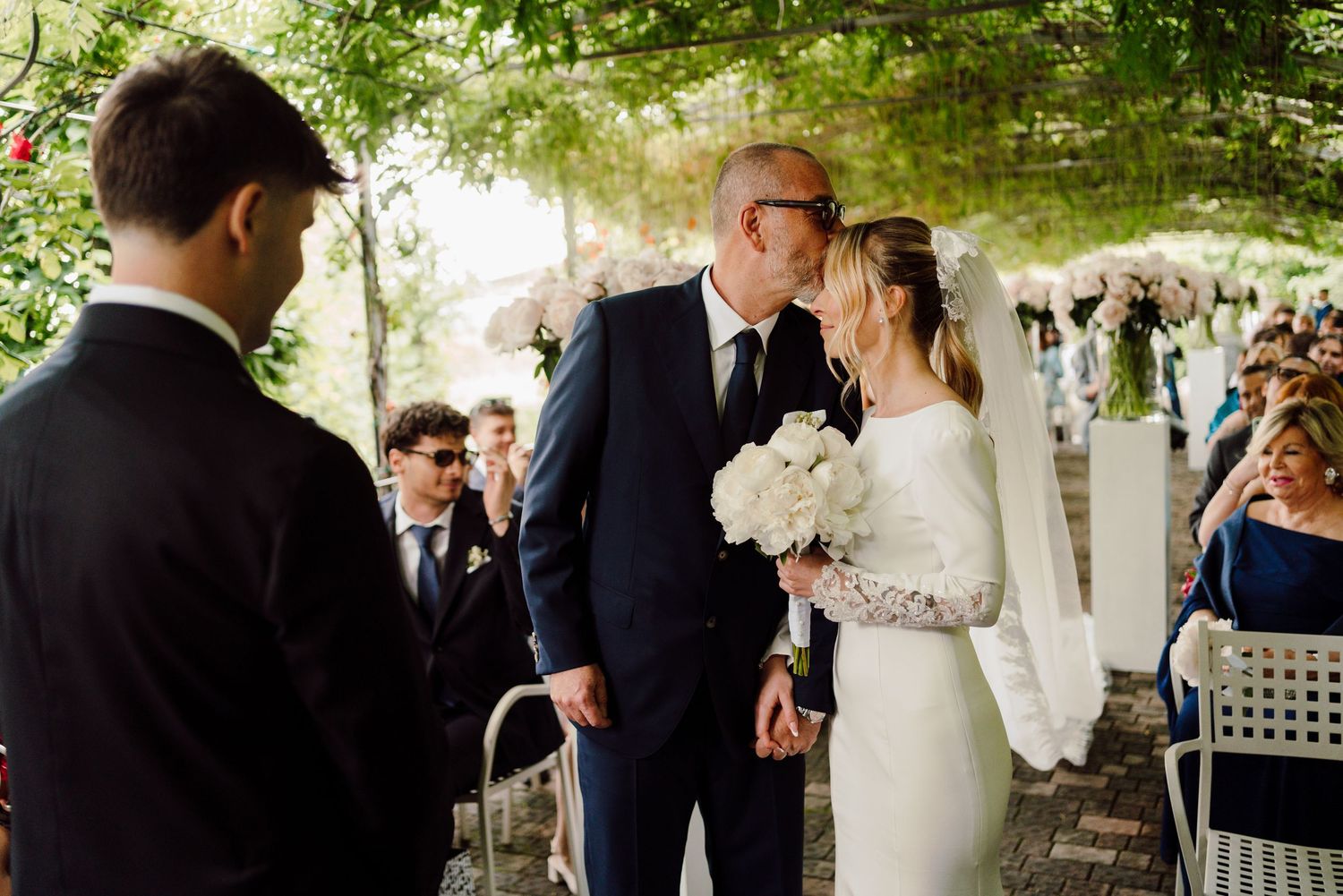 Father of the bride shares a farewell kiss with smiling bride holding white bouquet at altar.