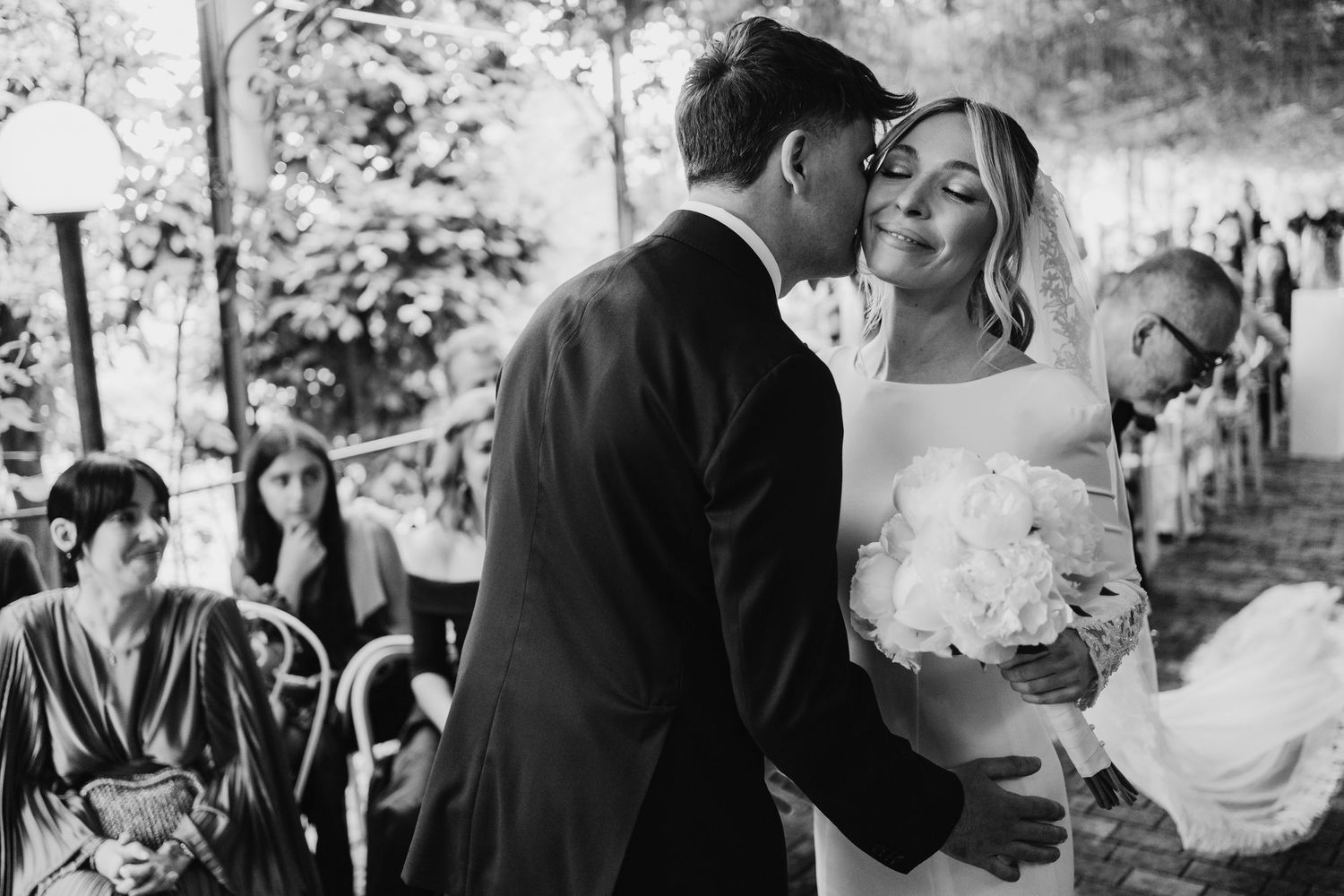 Groom tenderly kisses smiling bride holding white bouquet in black and white wedding photo.