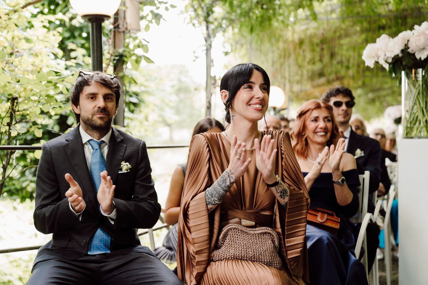Wedding guests clapping and smiling during an outdoor ceremony under lush green trees.
