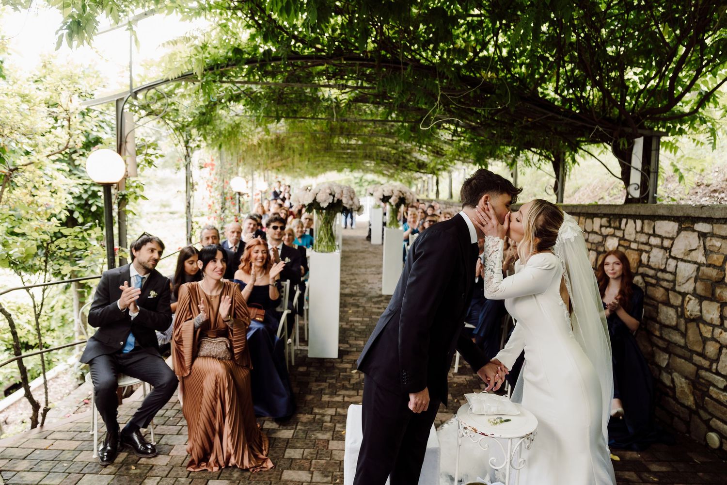 Newlyweds sharing a first kiss at their outdoor wedding ceremony while guests celebrate.