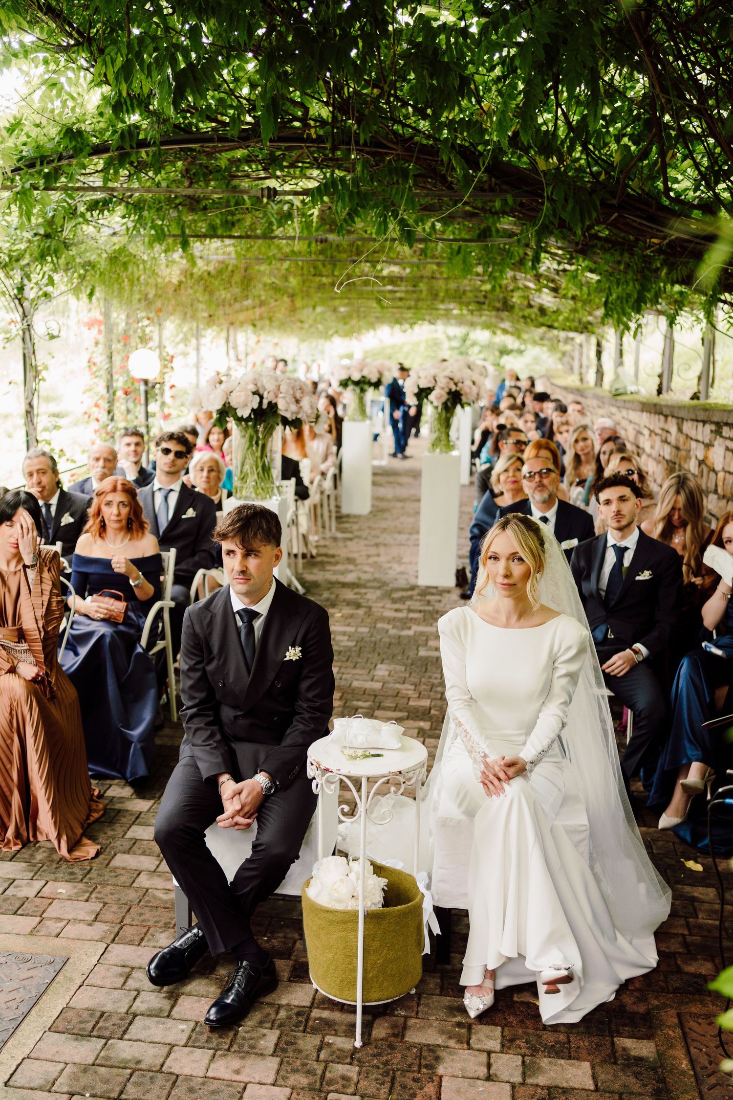 Bride and groom seated at outdoor wedding ceremony beneath a beautiful green vine canopy.