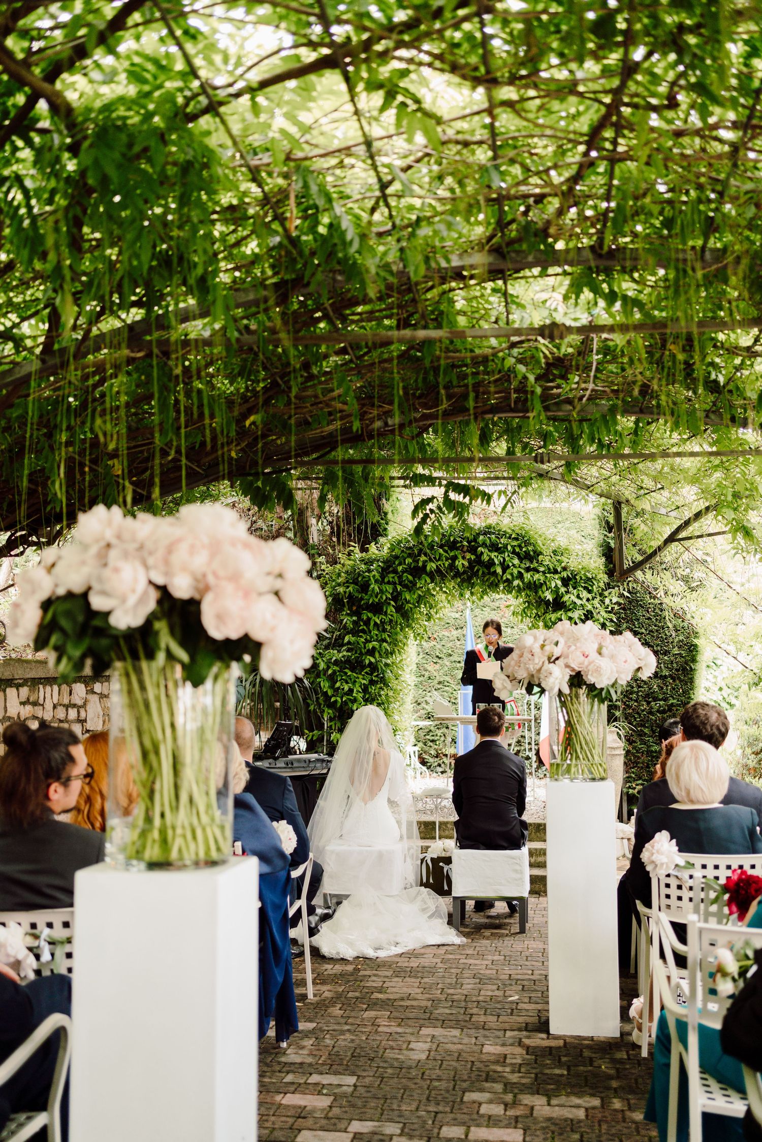 Elegant outdoor wedding ceremony under green canopy with white floral arrangements on pedestals.