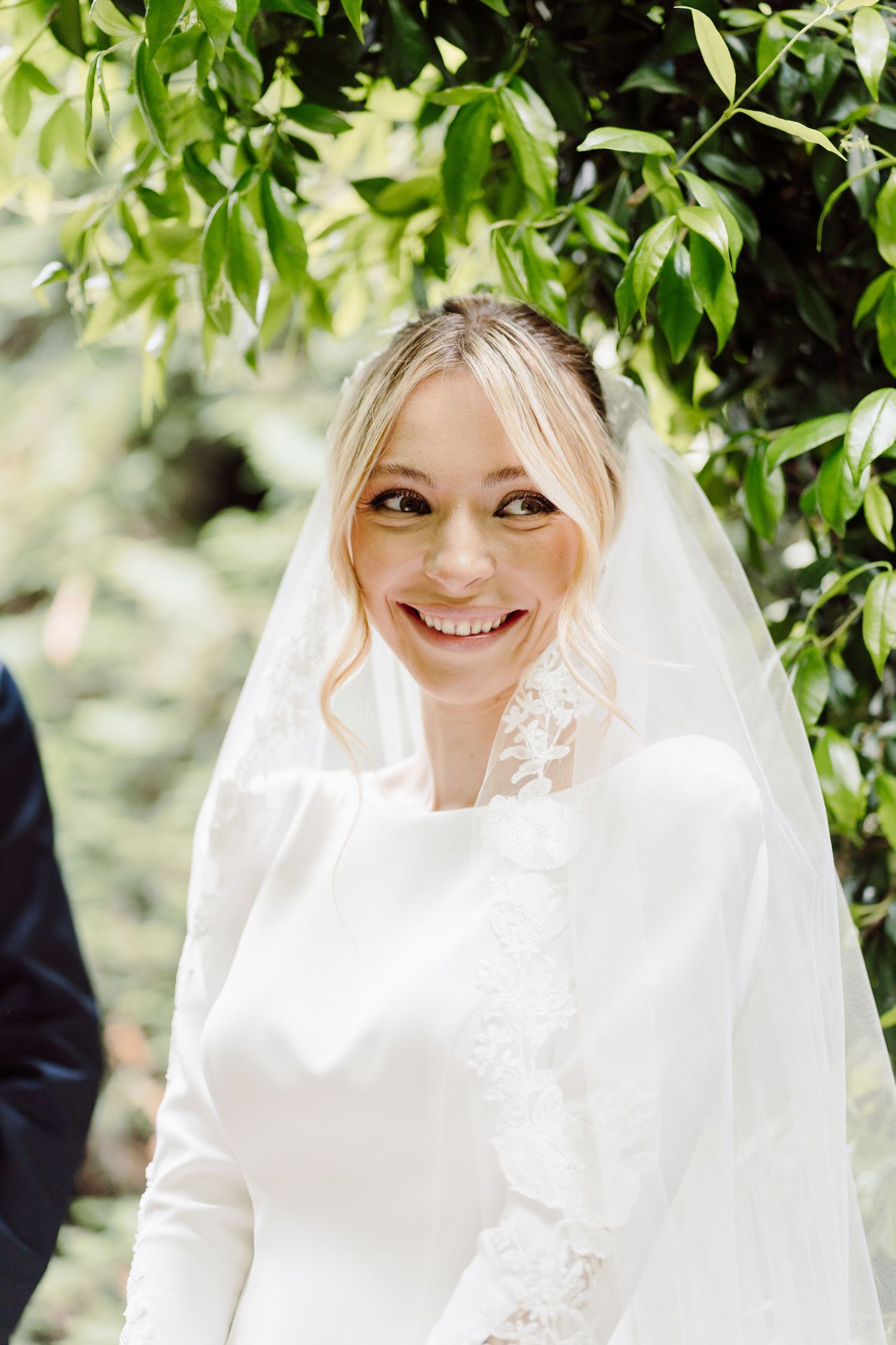 Beautiful smiling bride wearing lace veil and white gown surrounded by green foliage.