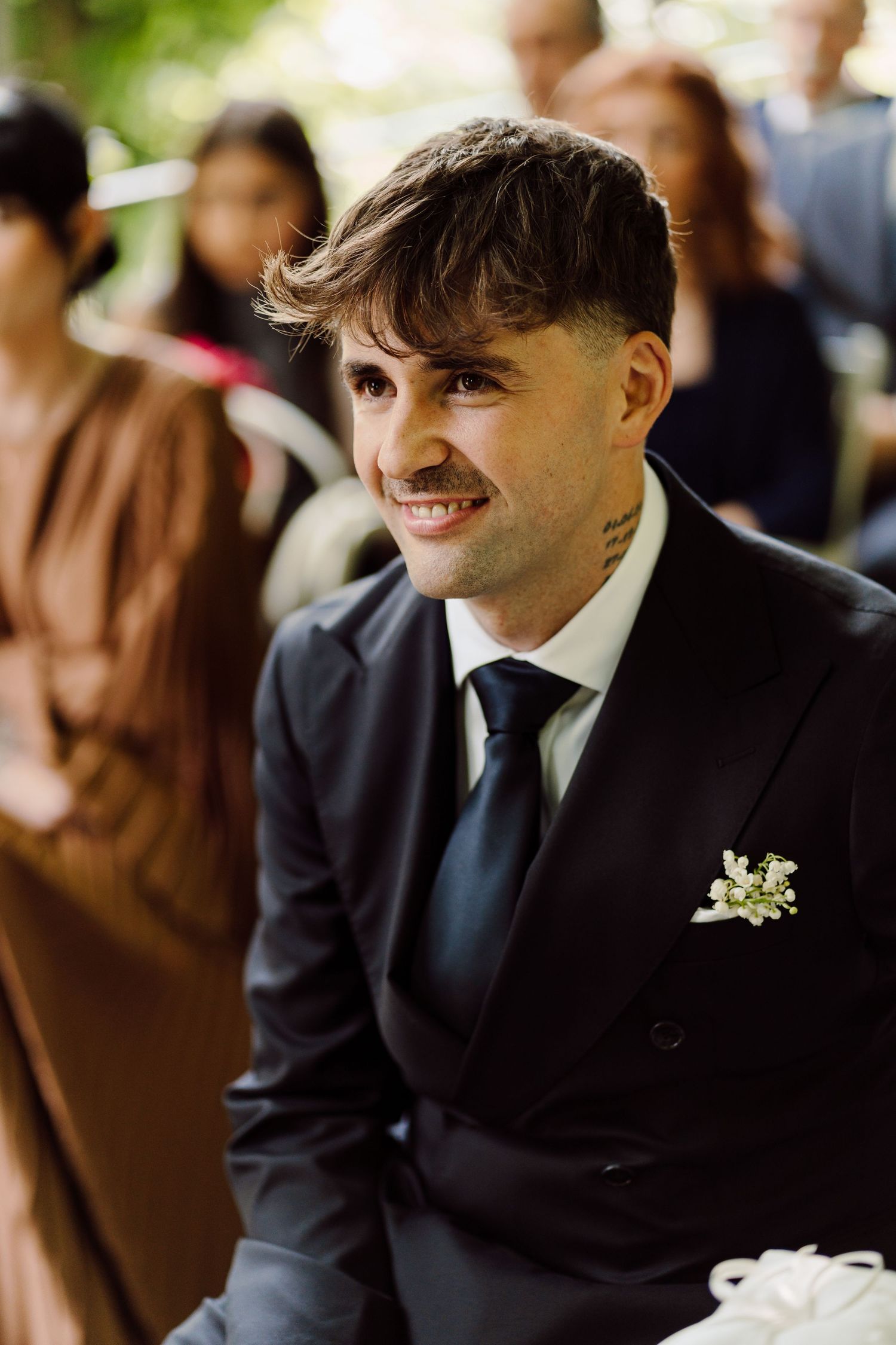 Smiling groom in dark suit with boutonniere seated at outdoor wedding ceremony.