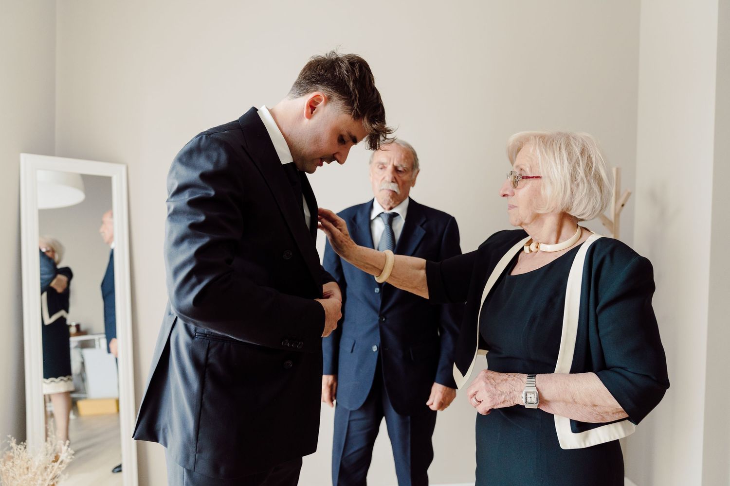 Groom in dark suit having tie adjusted by elderly woman with guests nearby.