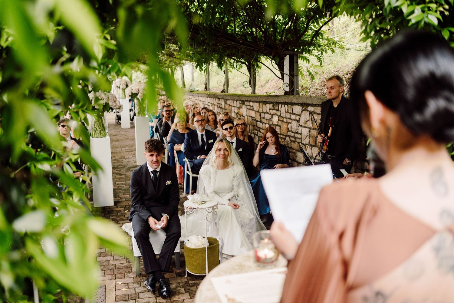 Wedding officiant reads vows as bride in white gown and groom in black suit sit during outdoor ceremony.