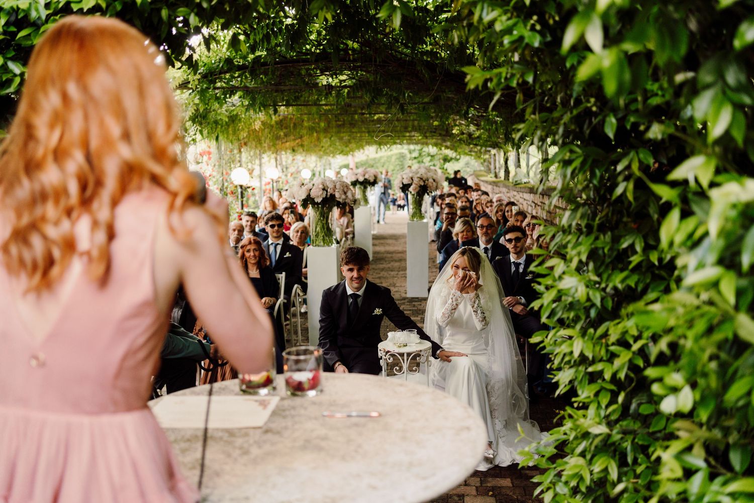 Outdoor wedding ceremony under green vine canopy with bride and groom at altar while bridesmaid speaks.