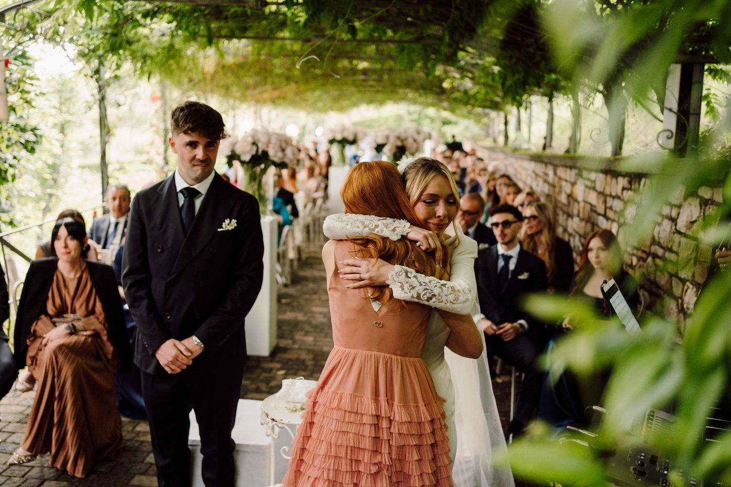 Bride in white wedding dress embraces bridesmaid in pink while groom watches during outdoor ceremony.