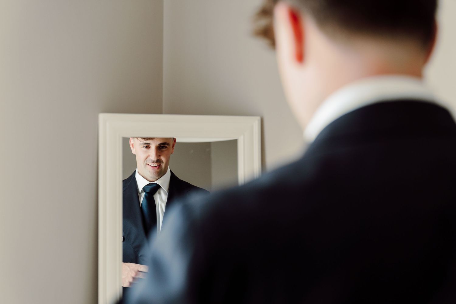Dapper groom in black suit and tie checking his appearance in a mirror.