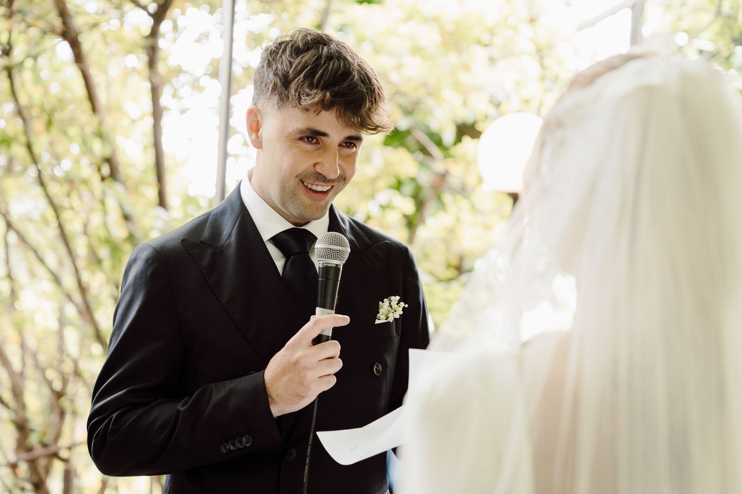 Smiling groom in black suit speaks heartfelt vows into microphone to veiled bride at outdoor wedding.