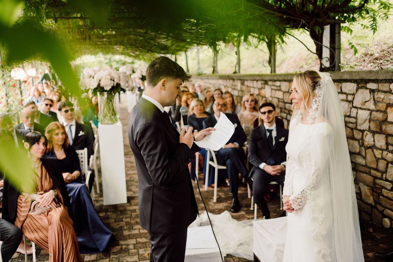Groom reads wedding vows to bride in white lace dress under green vine canopy with guests seated.