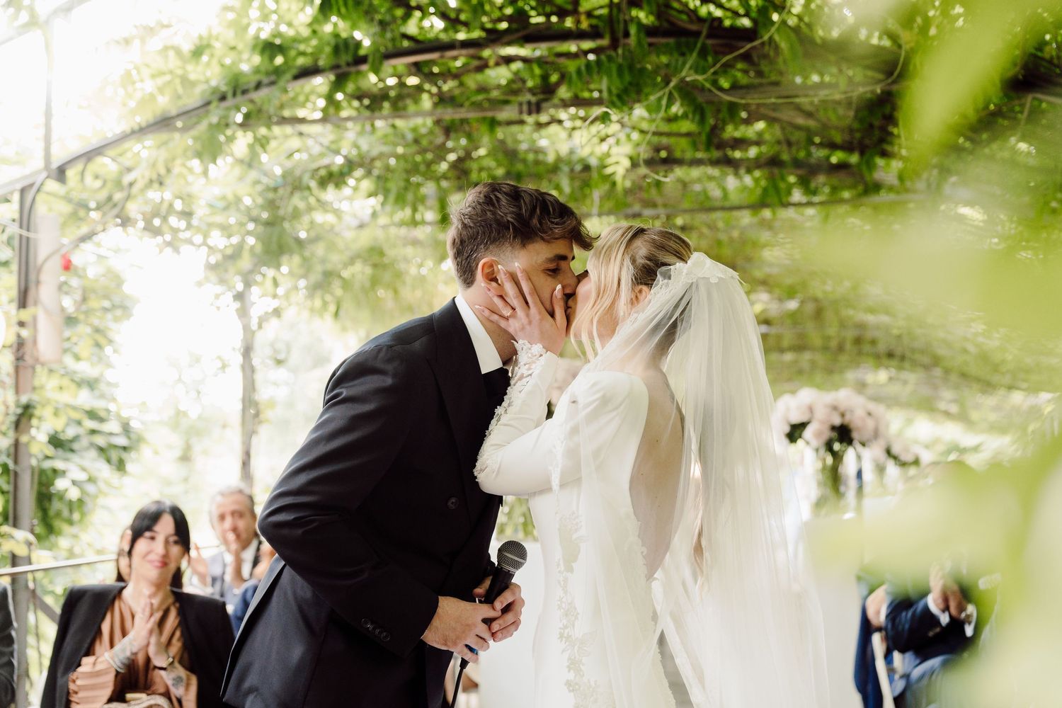 Bride and groom share first kiss under lush green vine canopy at outdoor wedding ceremony.