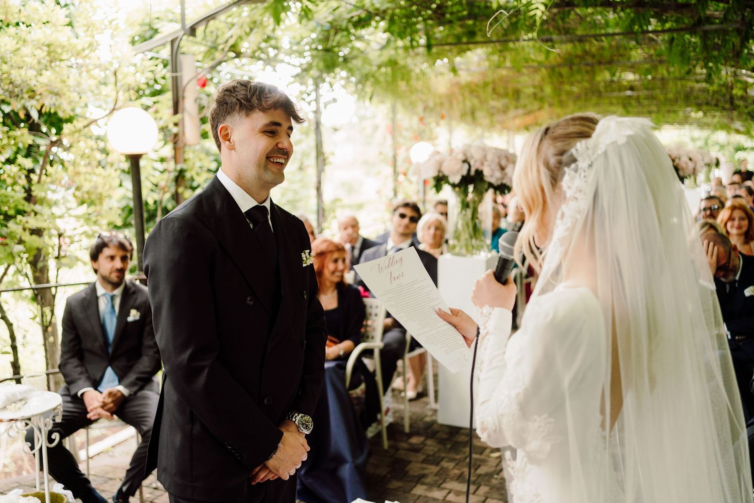 Groom smiles as veiled bride reads wedding vows during romantic outdoor ceremony with guests watching.