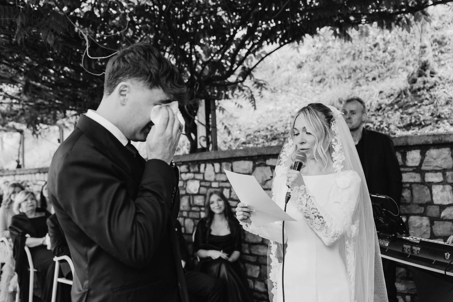 Emotional black and white photo of groom wiping tears as bride reads heartfelt vows at wedding.