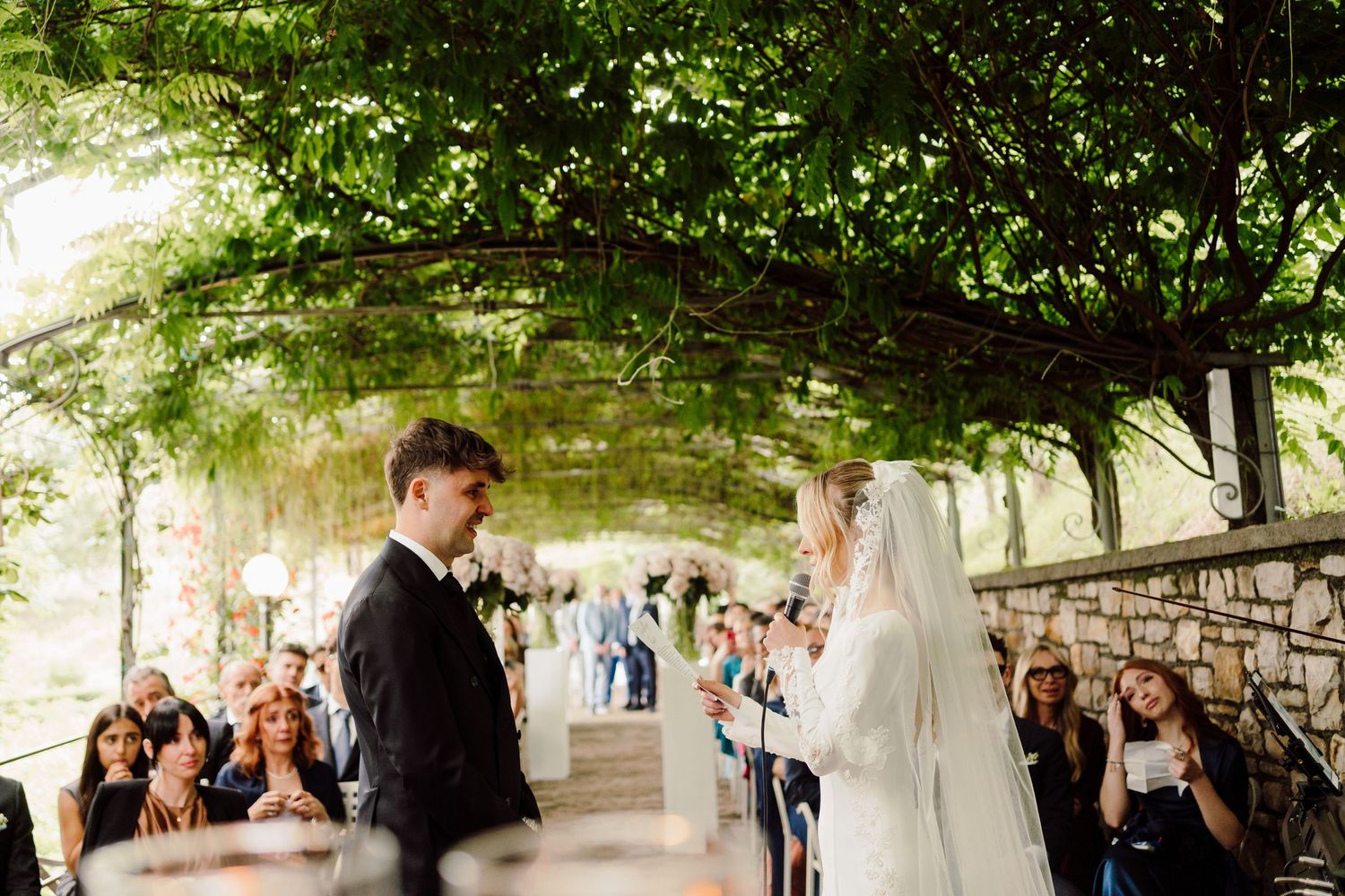 Bride and groom exchange vows under a lush green canopy with wedding guests seated along stone wall.