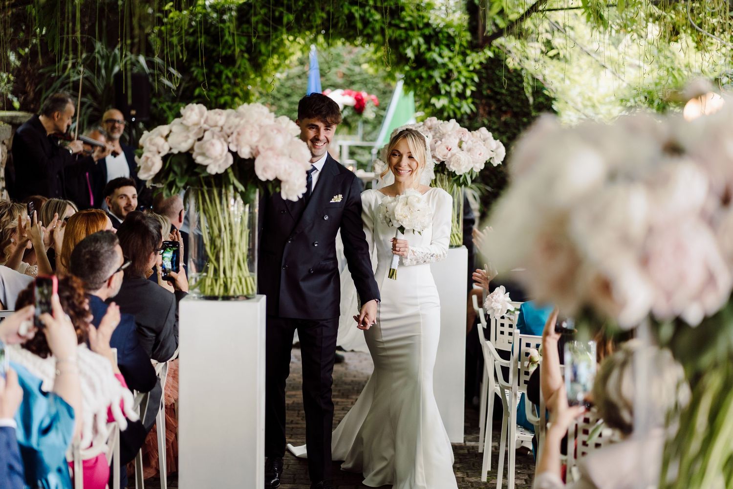 Bride and groom walk down the aisle hand in hand at an outdoor wedding ceremony with floral arrangements.