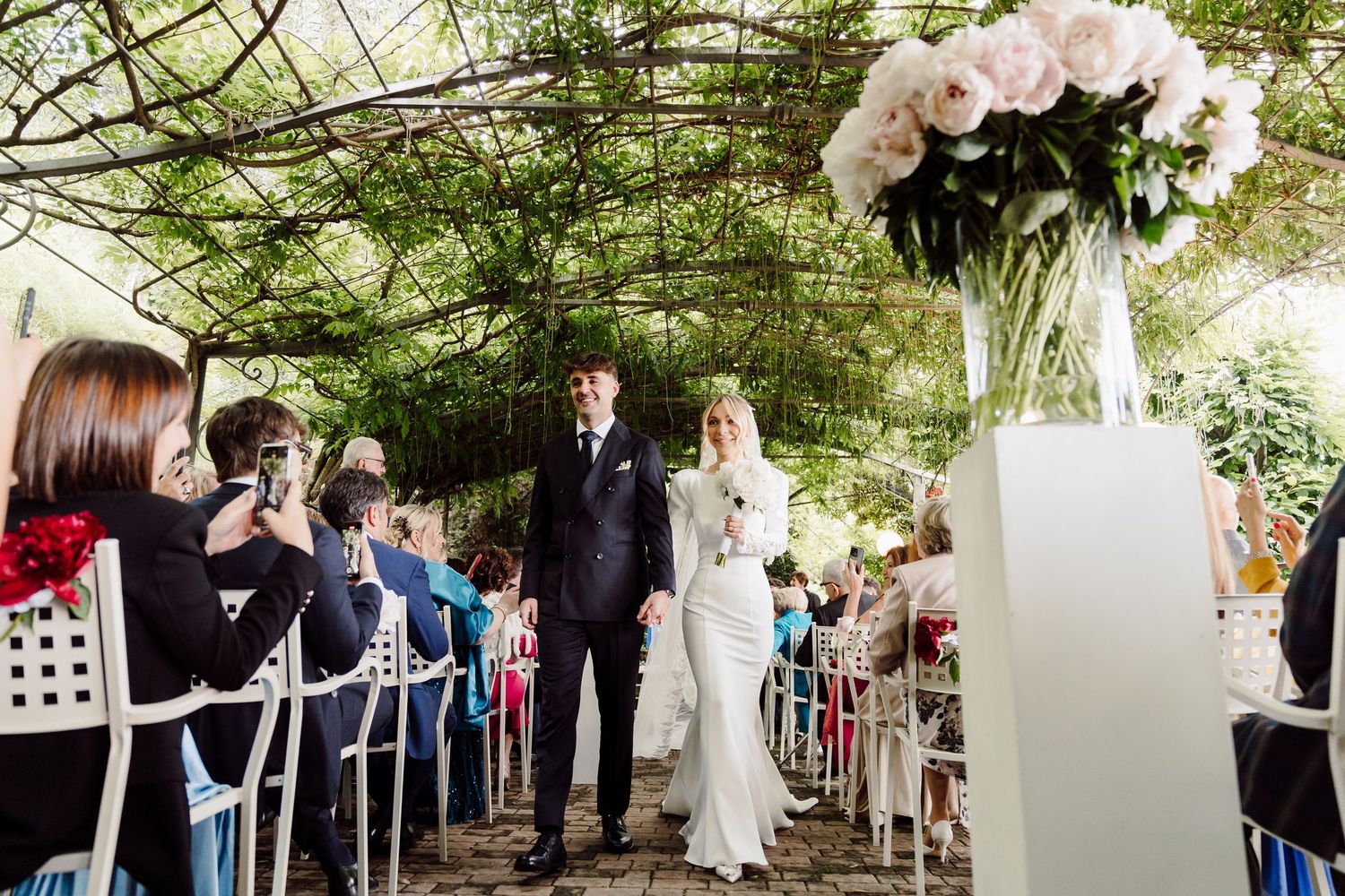 Newlyweds walk up the aisle under a lush green pergola surrounded by seated wedding guests.