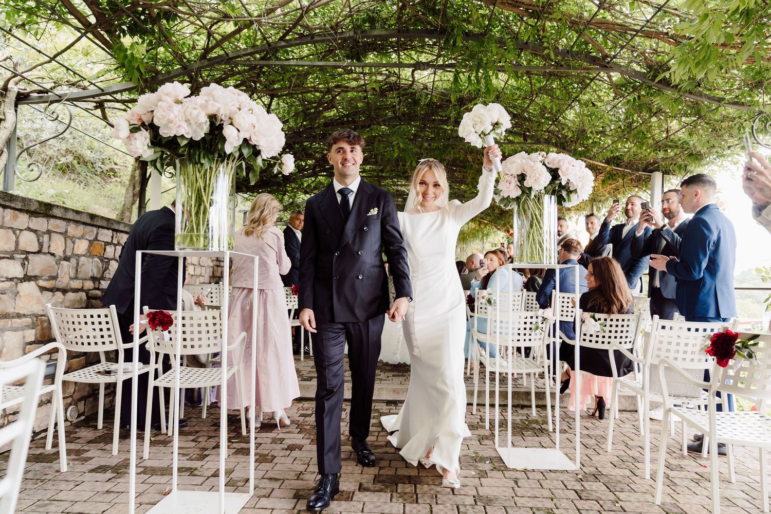 Joyful bride raises bouquet while walking with groom through elegant outdoor ceremony with white chairs.