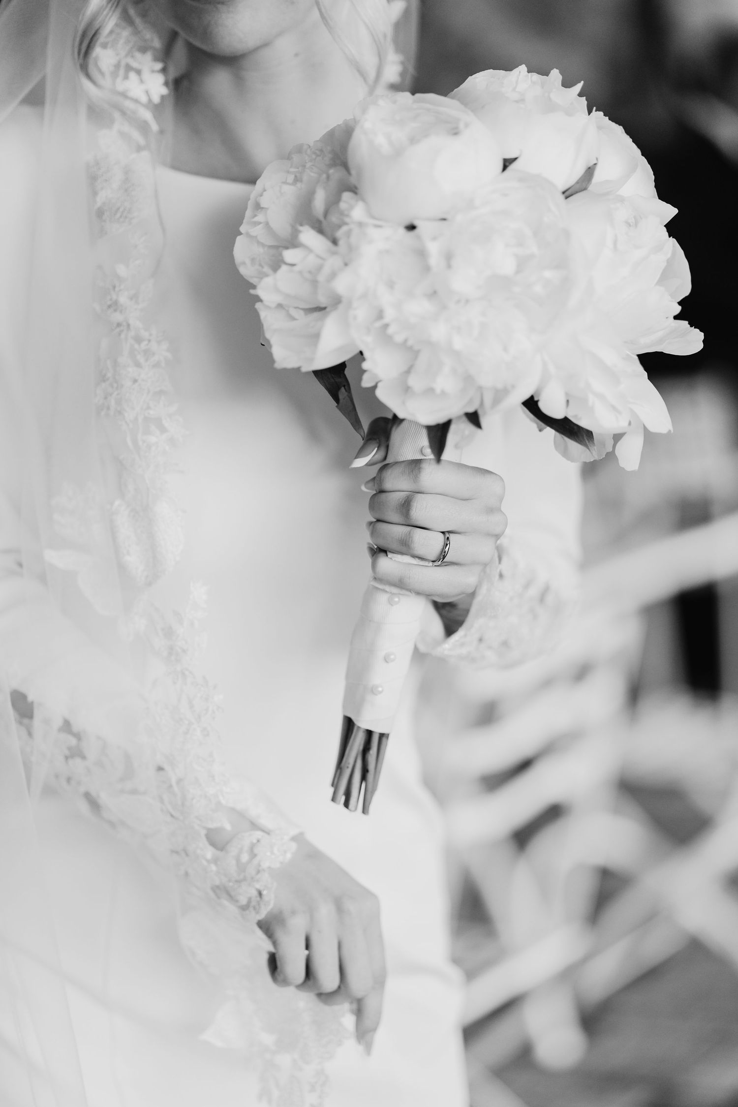 Black and white close-up of bride holding white peony bouquet with wedding ring visible on hand.