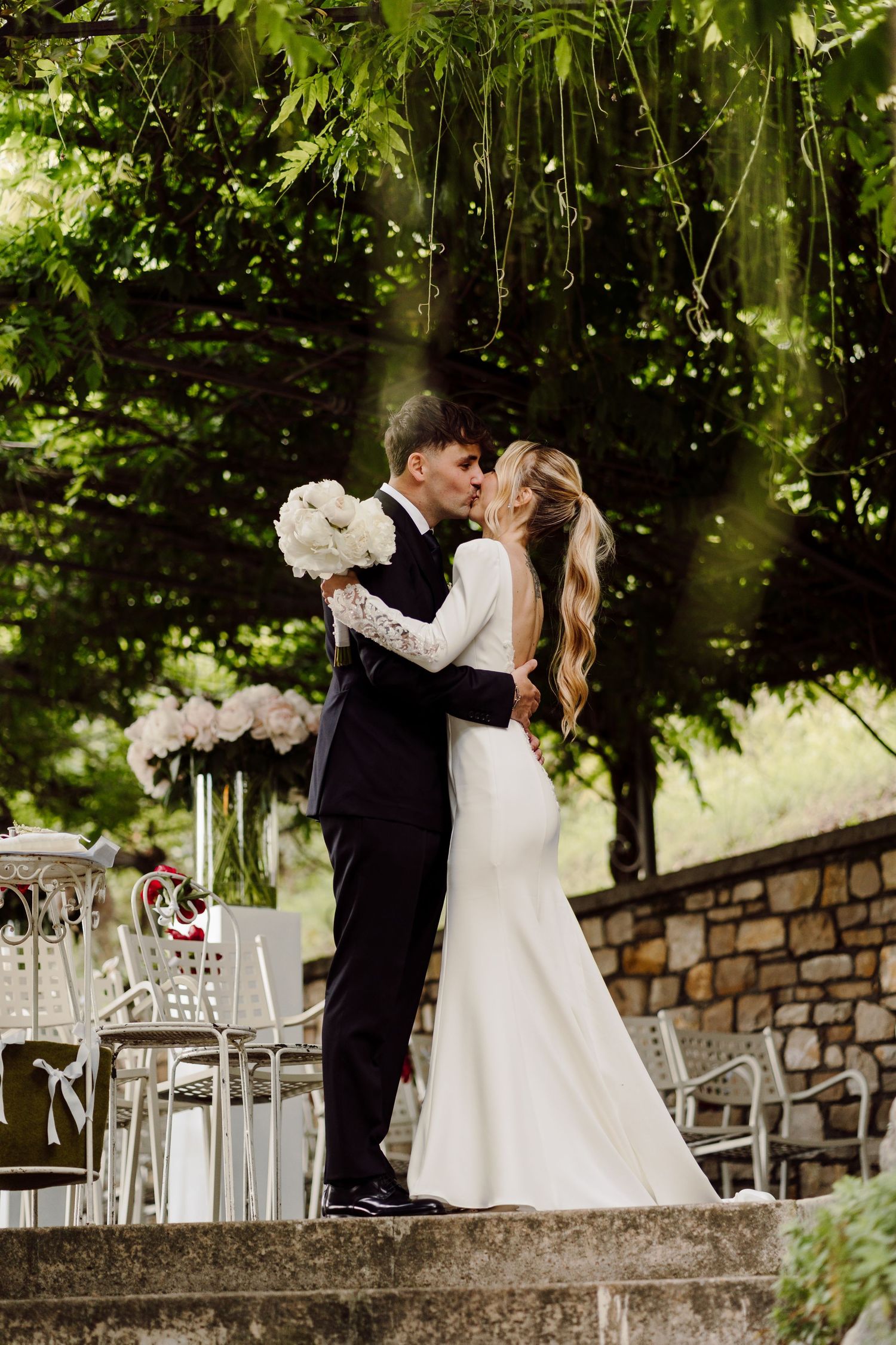 Bride and groom share first kiss under green foliage at an outdoor garden wedding venue.