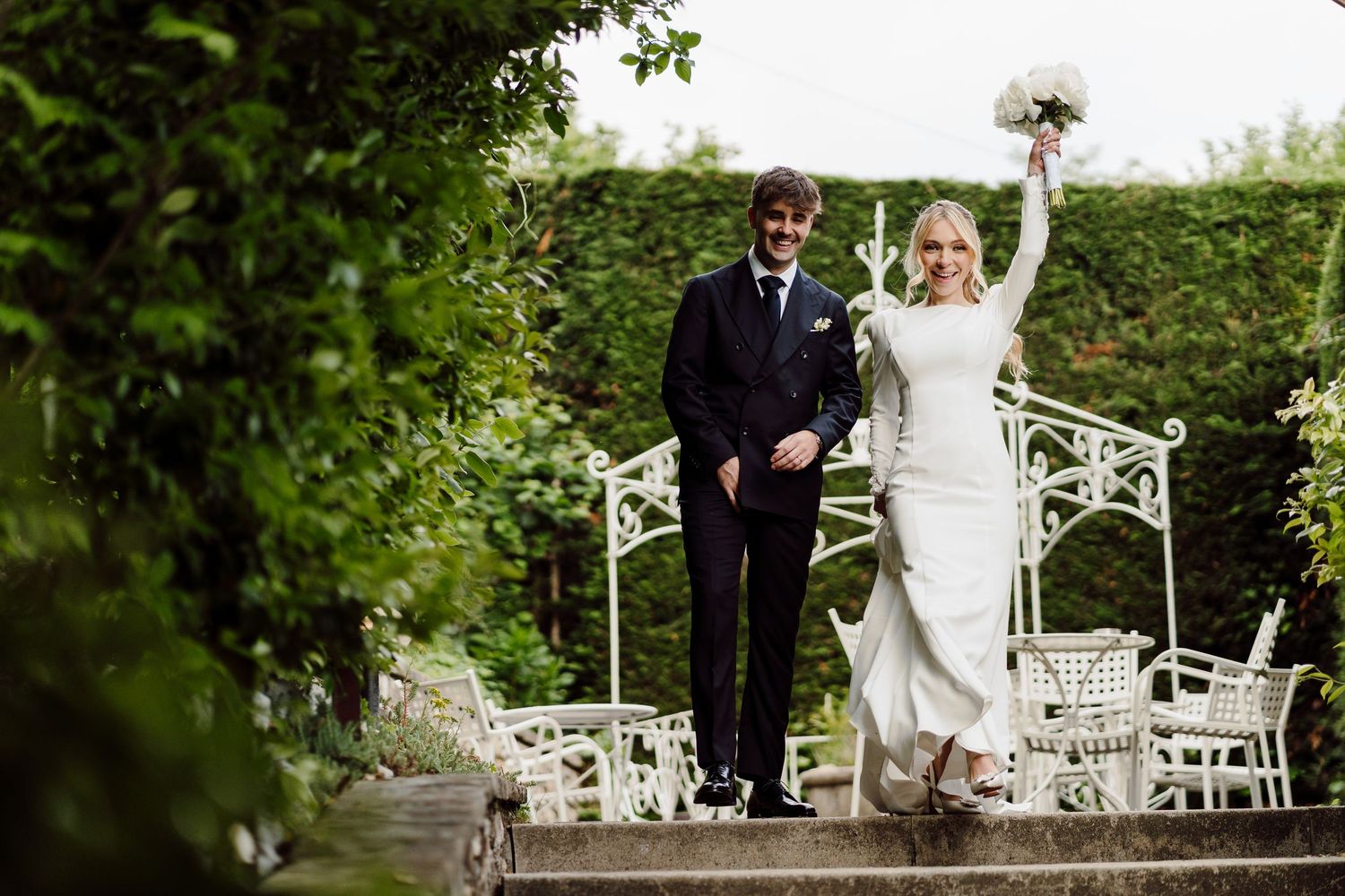 Smiling bride raises bouquet triumphantly while walking with groom through lush garden wedding venue.