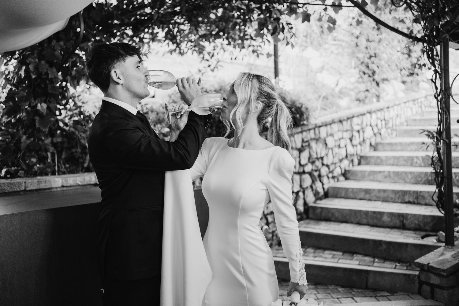 Black and white photo of groom drinking from bottle while bride laughs at outdoor wedding.