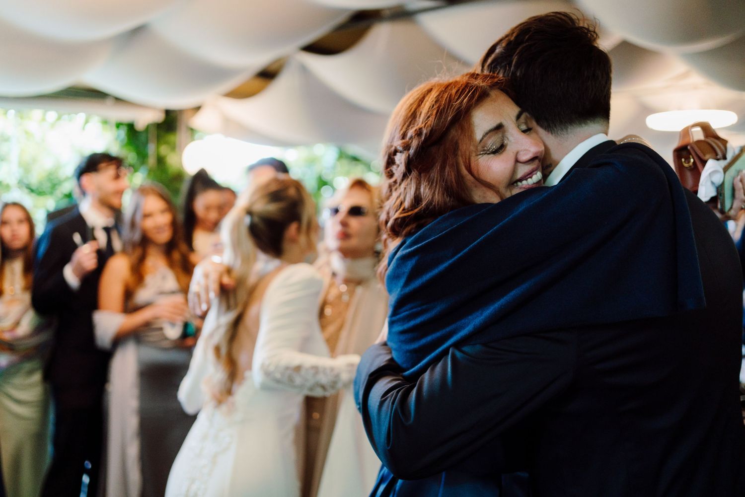 Emotional groom embraces red-haired guest during wedding reception while guests celebrate in background.