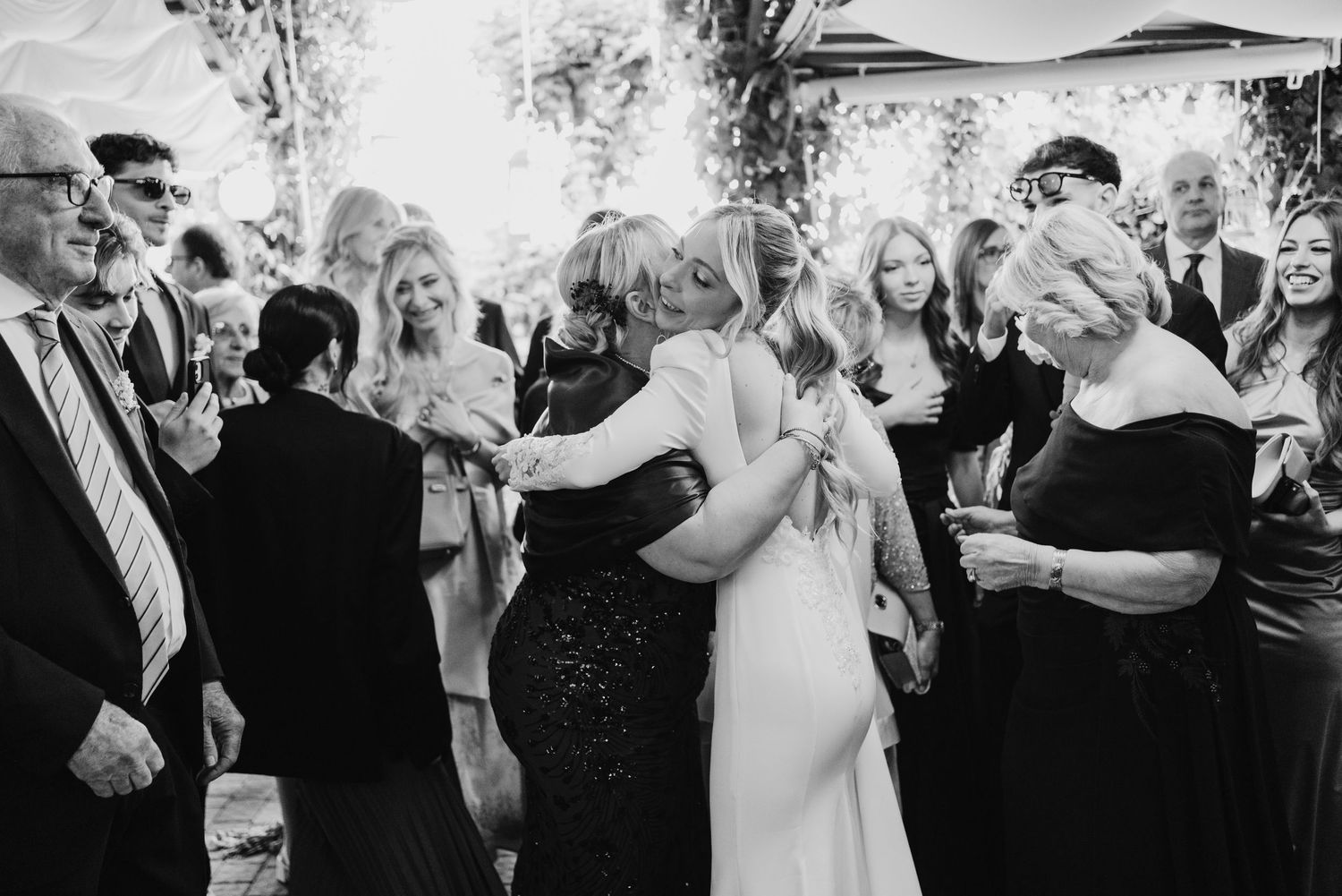 Black and white photo of bride embracing guest warmly surrounded by smiling wedding attendees.