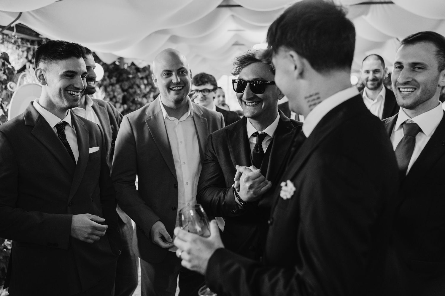 Black and white photo of groomsmen laughing and socializing at a wedding reception under a tent.