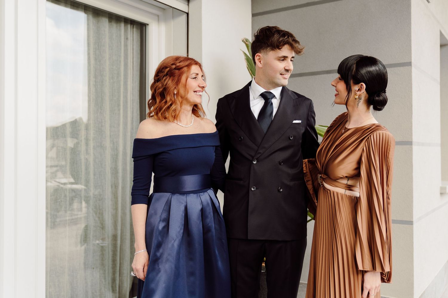 Groom in black suit posing with two elegantly dressed women on a balcony.