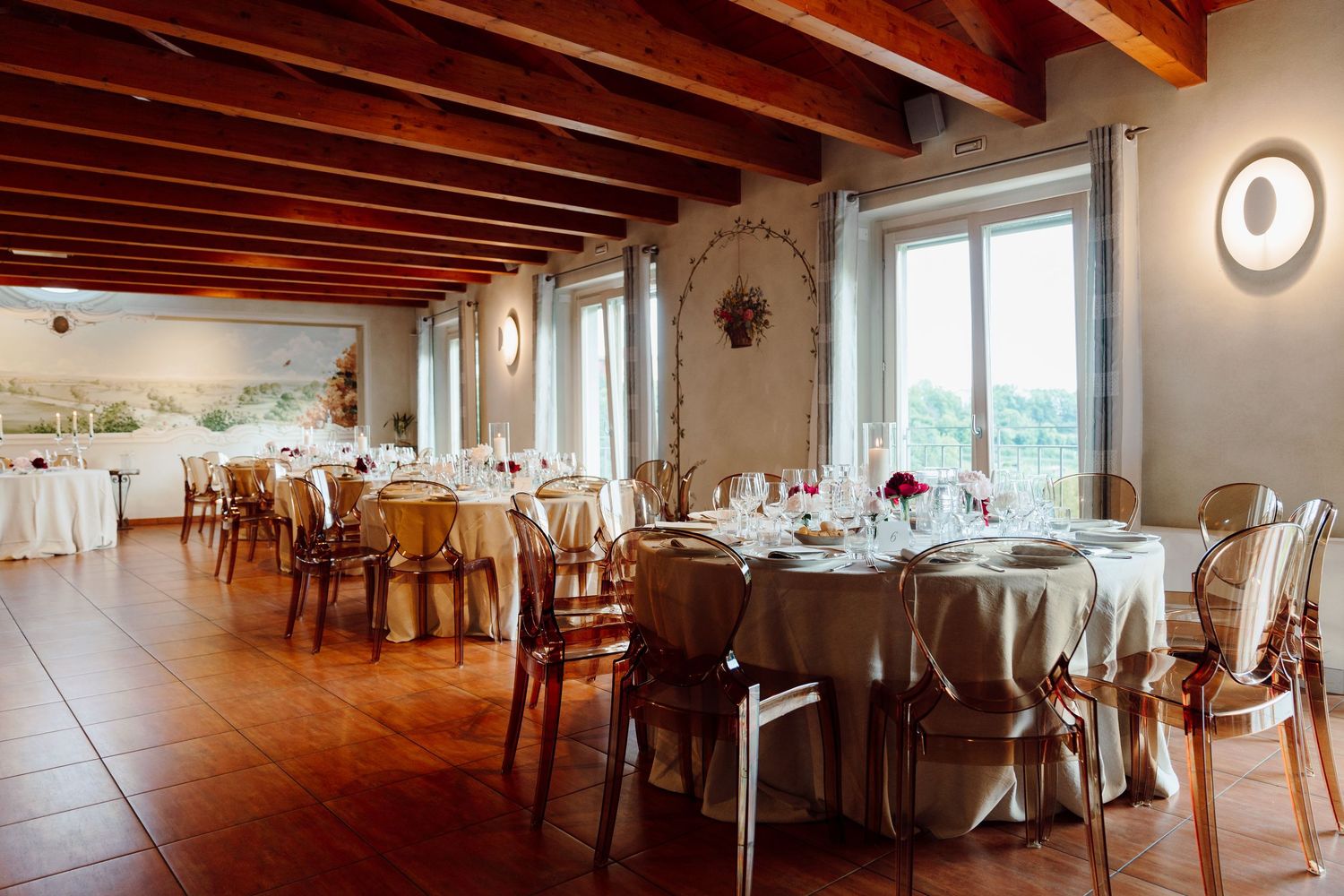 Rustic wedding reception hall with wooden beam ceiling, round tables, and ghost chairs.