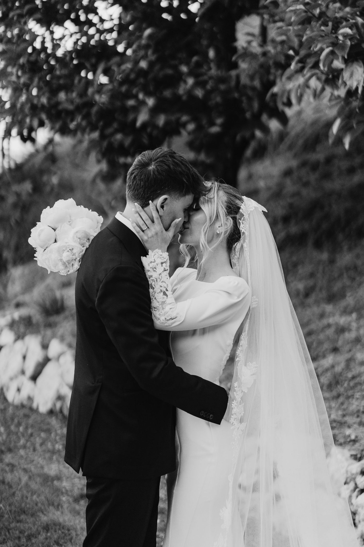 Black and white portrait of bride and groom sharing an intimate moment outdoors under a tree.