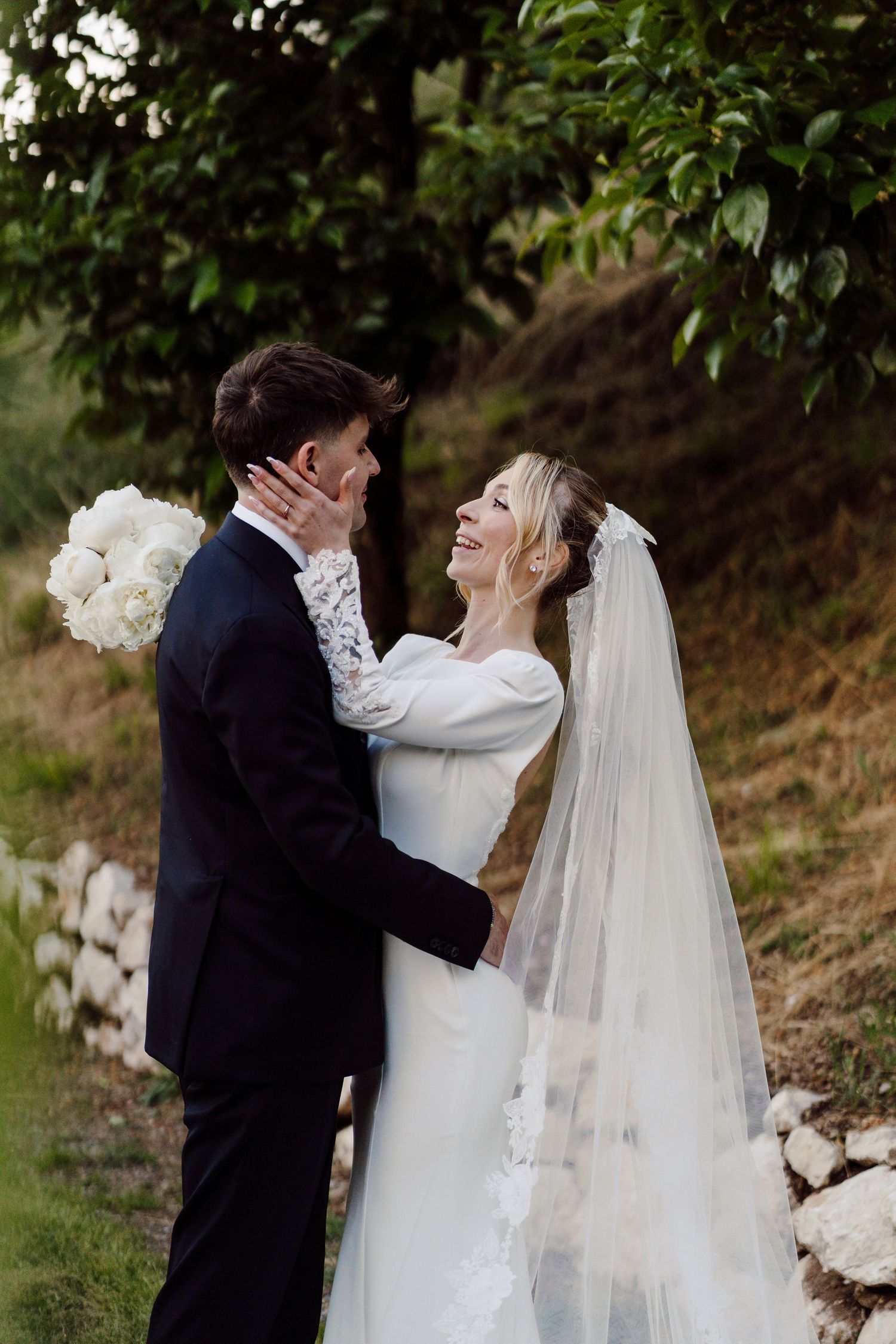 Bride laughing joyfully while holding grooms face during outdoor wedding portrait in lush greenery.