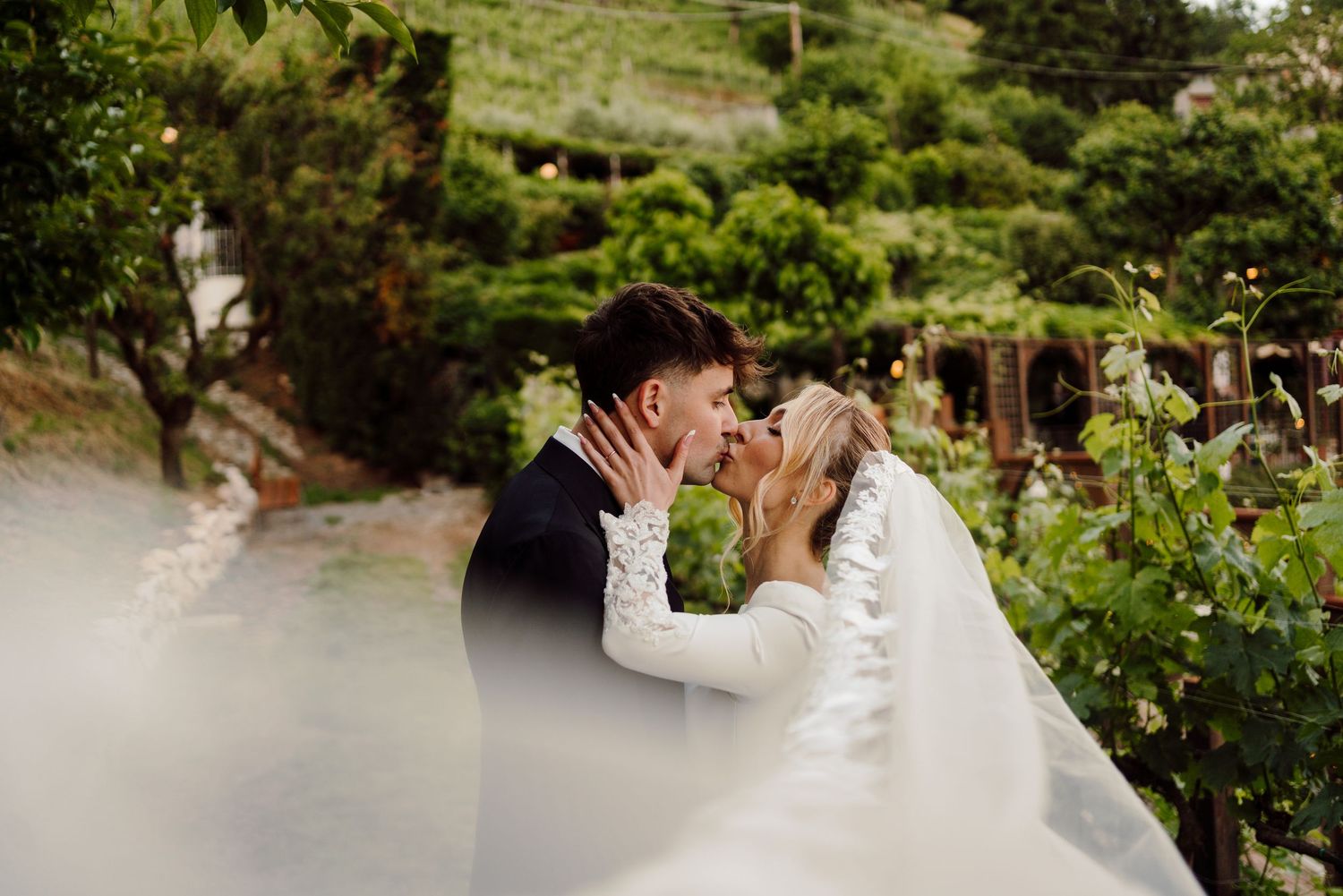 Couple kissing passionately among green vineyard vines, bride in long-sleeved white gown with flowing veil.